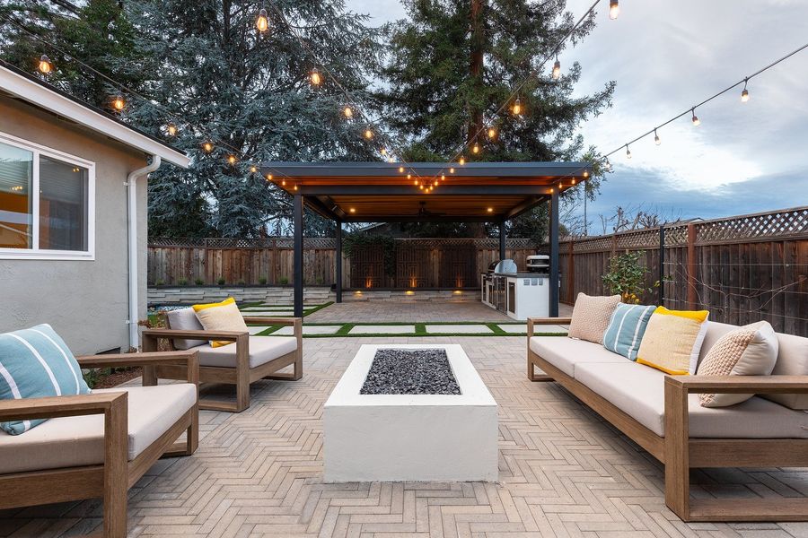 Herringbone paver patio with a white square fire table, wood-framed sofas and chairs, and a steel pavilion with string lights beyond