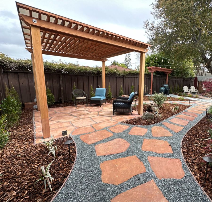 Wood pergola over a terracotta flagstone patio with wicker armchairs, a flagstone path curving through gravel and mulch beds, and a second small pergola in the distance.
