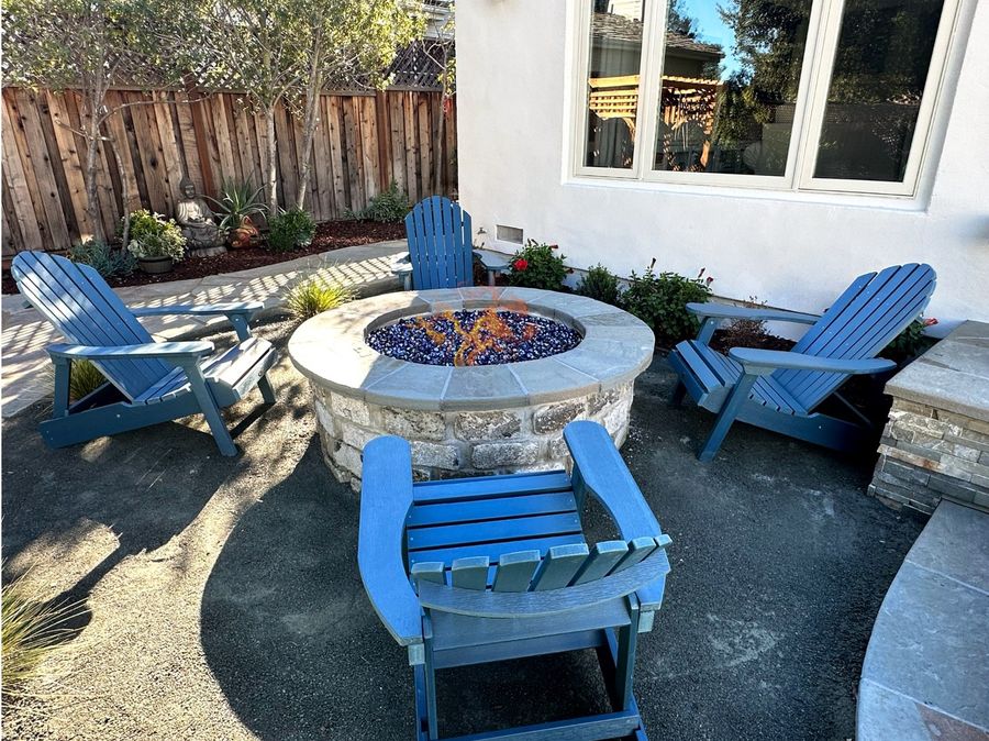 Round stone fire pit with a bluestone cap and blue fire glass, surrounded by four blue Adirondack chairs on dark gravel, with a stacked stone wall and flagstone path beside a white house.