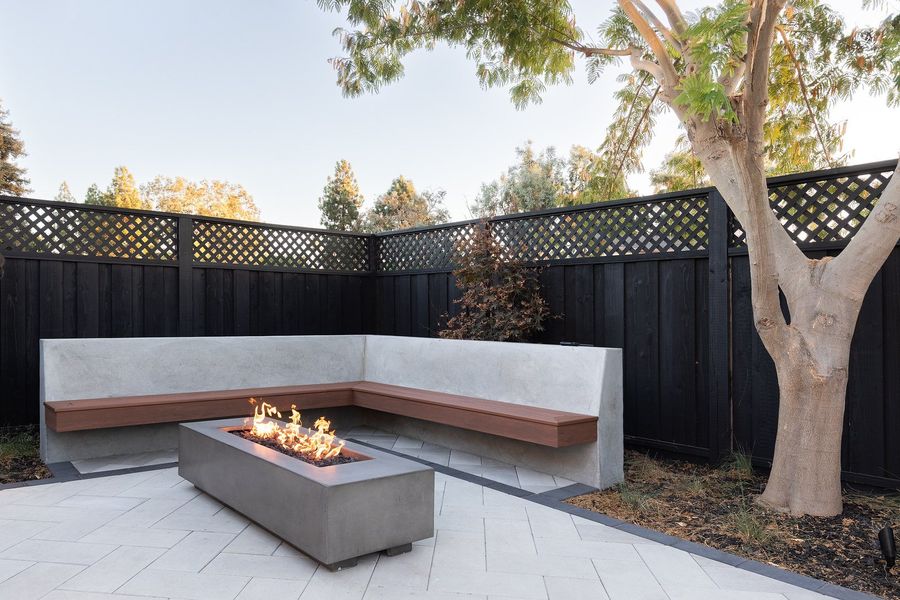 Concrete corner bench with a wood seat and a linear fire table on gray pavers beside a mature tree