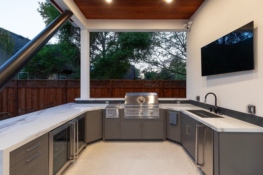 U-shaped outdoor kitchen with a stainless grill, side burner, sink and glass-front refrigerators under a timber ceiling