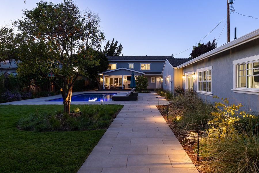 Dusk view down a straight slab path beside beds of ornamental grasses and lit shrubs, with a lawn and an orange tree to the left