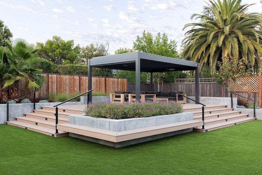Wide raised deck with a staircase, concrete planter filled with lavender and a louvered pergola over lounge seating at dusk
