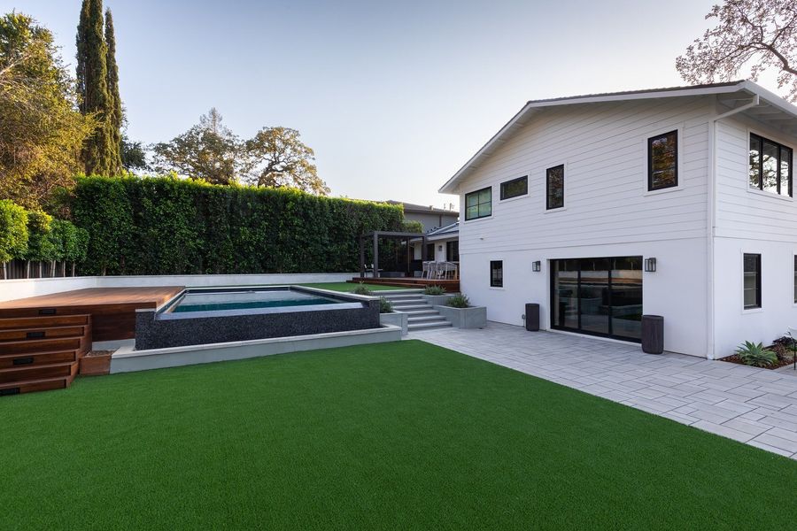 White house with black windows, a paver walk, lawn and a raised hardwood deck with steps in the background