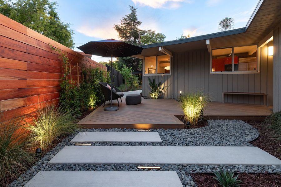 Corner of the entry deck at golden hour with a black umbrella, lounge chair, climbing plants on a wood screen and a stepping-pad path