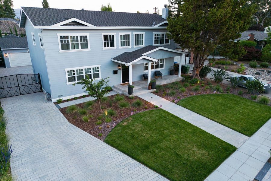Elevated view of a brick-pattern paver driveway beside the front lawn, with a black iron gate at the left and planted beds along the street edge