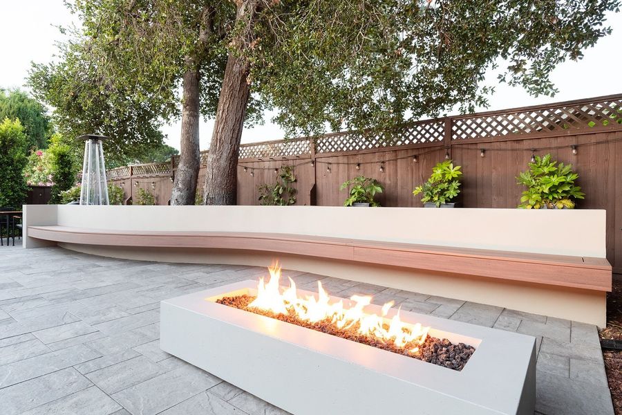 Linear gas fire table burning in front of a curved white bench with a wood seat on gray pavers