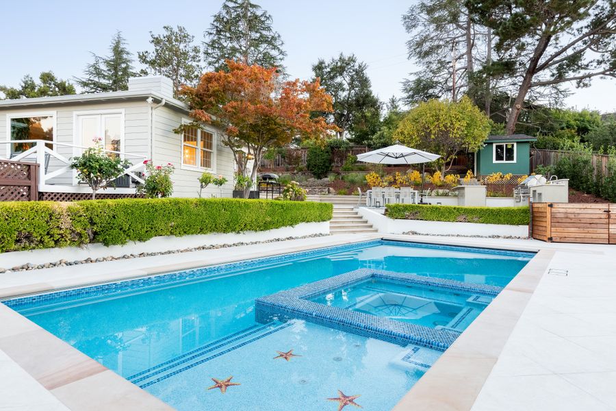 Pool with an integrated raised spa in a pale stone surround, hedge and house behind