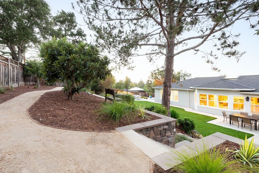 Curving gravel path around a mulched bed with a small tree, a stone retaining wall and the lit house below