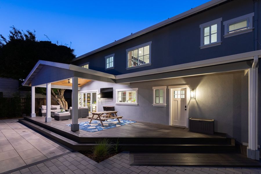 Blue-hour view of the covered deck raised above pale slab paving, with white posts, a gabled roof and a white door beside the lit patio