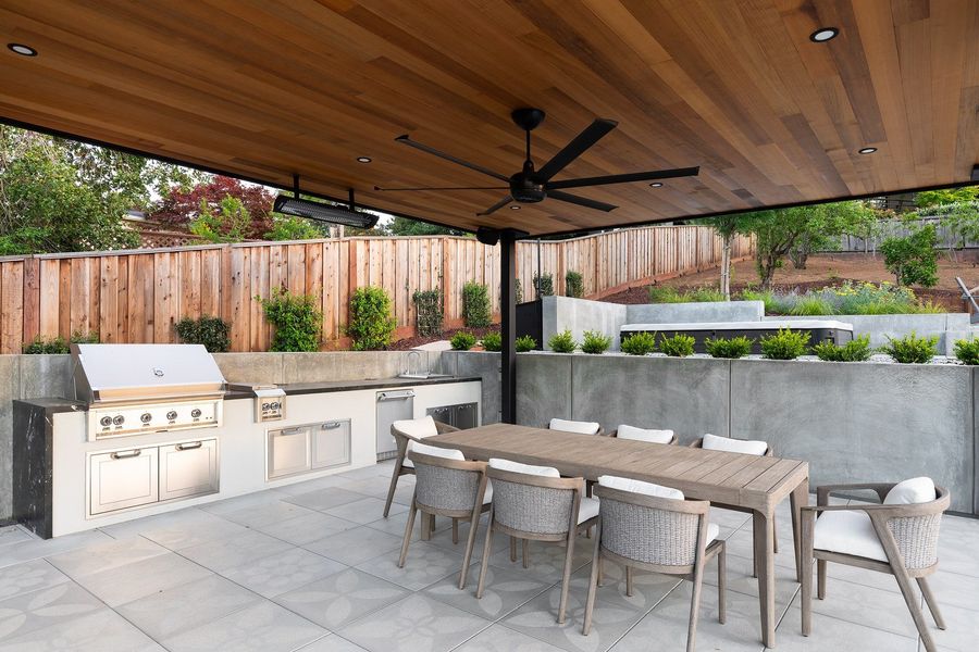 Close view under the pavilion ceiling: grill, prep counter, ceiling fan, heater and a dining table with woven chairs