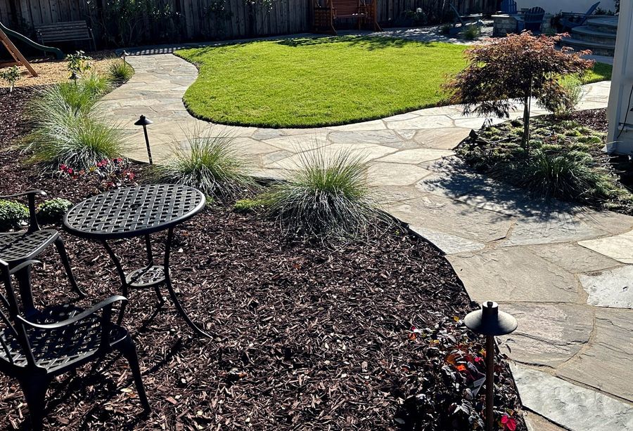 Curved flagstone path around a rounded green lawn, with blue-green ornamental grasses, a small red-leaf maple, a bistro table on mulch and path lights.
