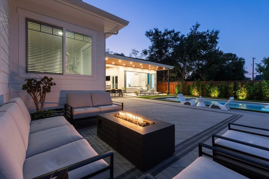 Low view past the fire table and sofas to the lit cabana across the paved yard at dusk
