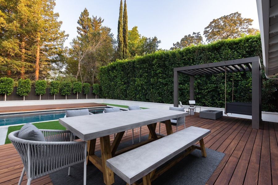 Long concrete dining table and bench on a hardwood deck with a dark pergola swing at the far end and a clipped hedge behind