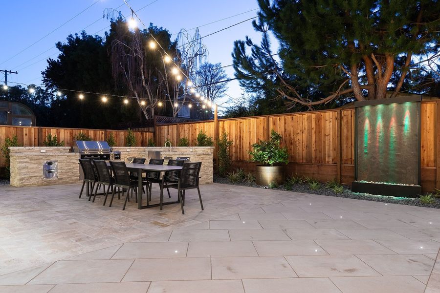Dusk view across the paved patio to a freestanding water wall in the fence and the kitchen island with dining table under string lights