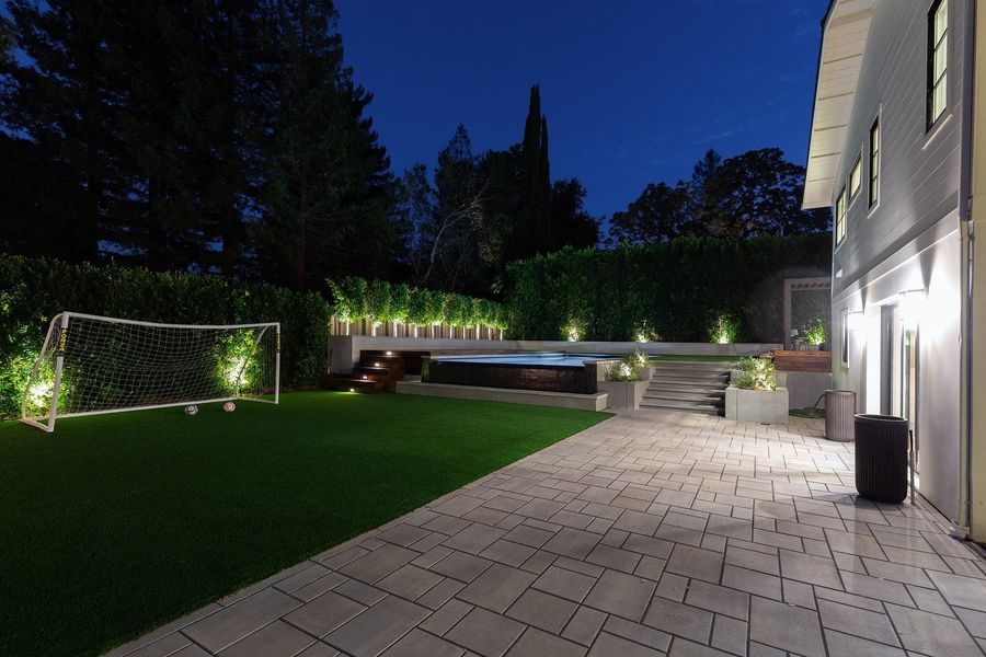 Night view of a grey paver terrace leading to a lit hedge, a soccer goal and a stepped wall