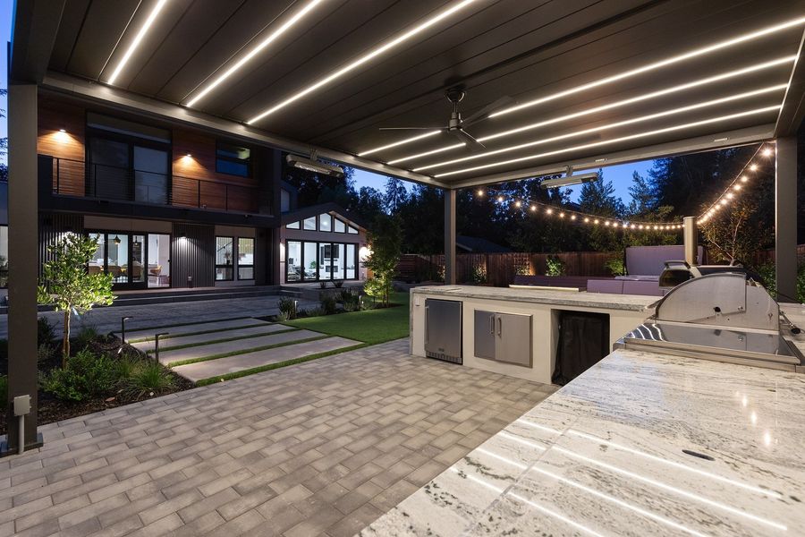 Close view across the stone counter and stainless fridge to string lights and the lit house across the terrace