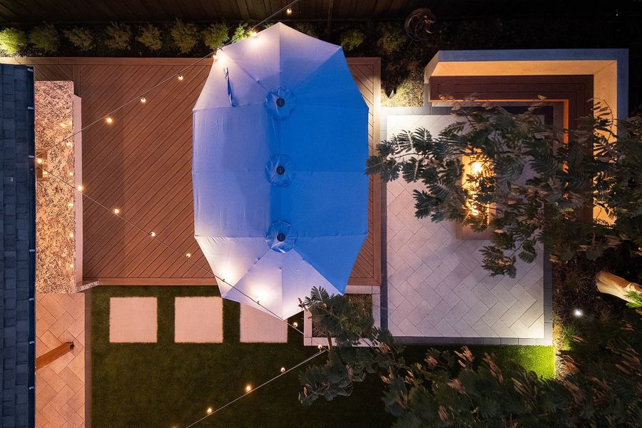 Aerial night view of a wood-look deck, a granite kitchen counter, stepping pavers in turf and a lit fire court with a corner bench