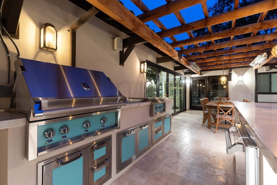 The same outdoor kitchen at night with warm wall lanterns, stainless grill and dark blue sky through the pergola beams