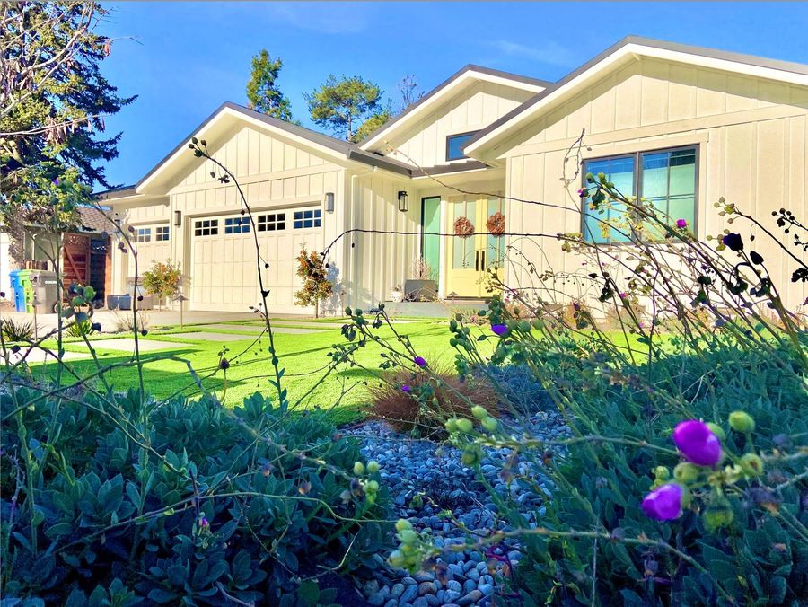 Low front bed of blue-green succulents with purple flower buds and river rock, with a bright green lawn, stepping pavers and a white house with a garage and yellow door behind.