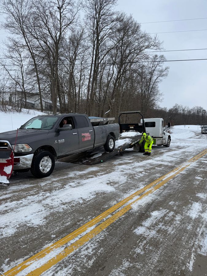 Gray pickup with a plow blade and a person in a yellow safety jacket beside a flatbed truck on a snowy road