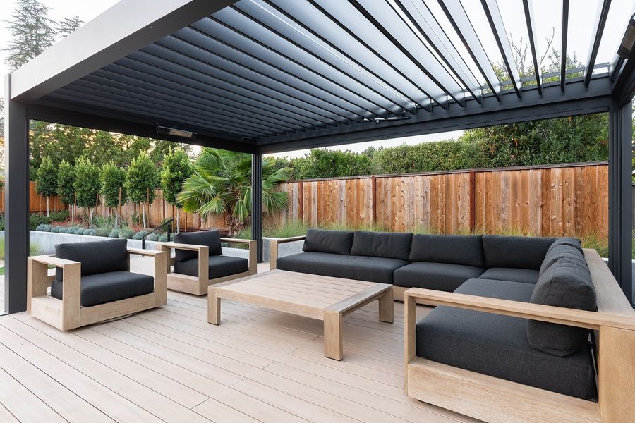 Close view under a black louvered pergola with an L-shaped sofa, armchairs and wooden coffee table on a pale deck