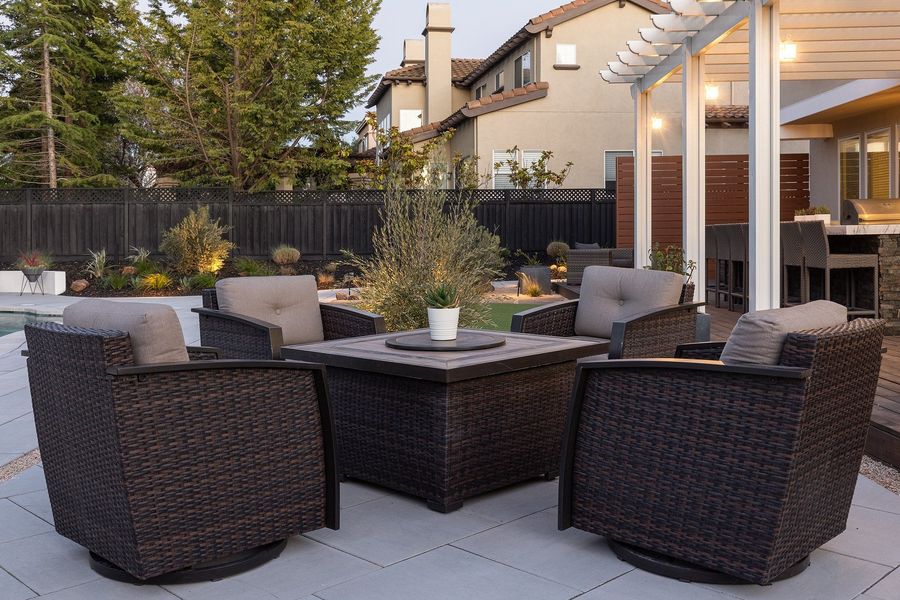 Dusk view of woven armchairs around a fire table on pale paving, with a white pergola and grasses behind