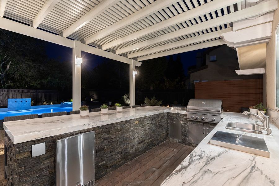 Night view of the stacked-stone kitchen island with veined white counters, a built-in grill, sink and refrigerator under the white louvered pergola