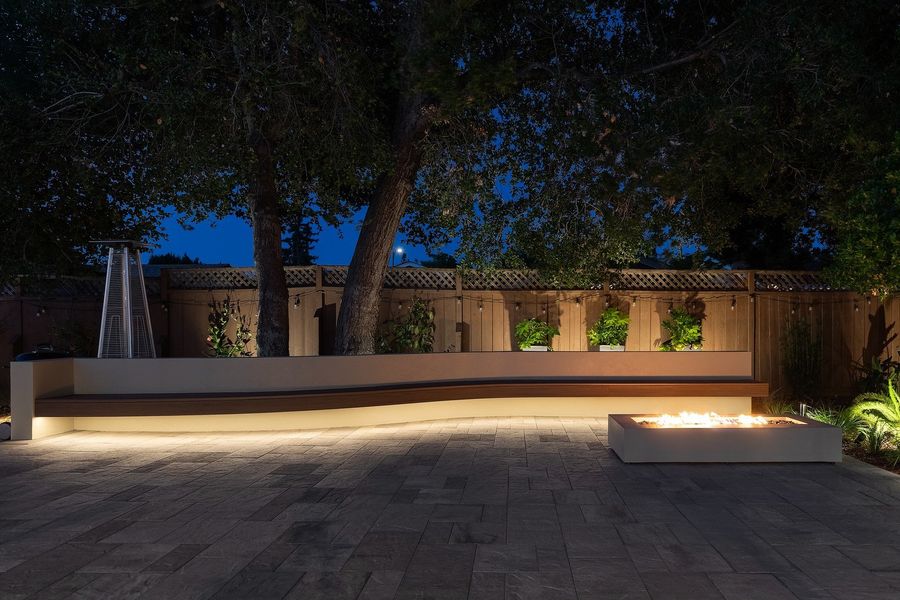 Night view of the curved bench lit from below, with the fire table burning on dark pavers and string lights along the fence
