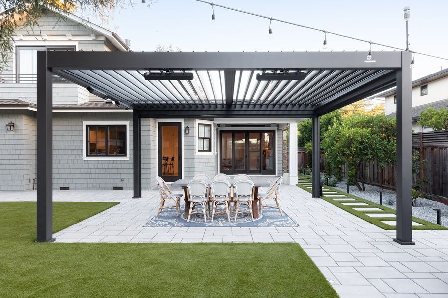 Louvered steel pergola with overhead heaters over a dining table on pale pavers, with stepping pads and turf beside it
