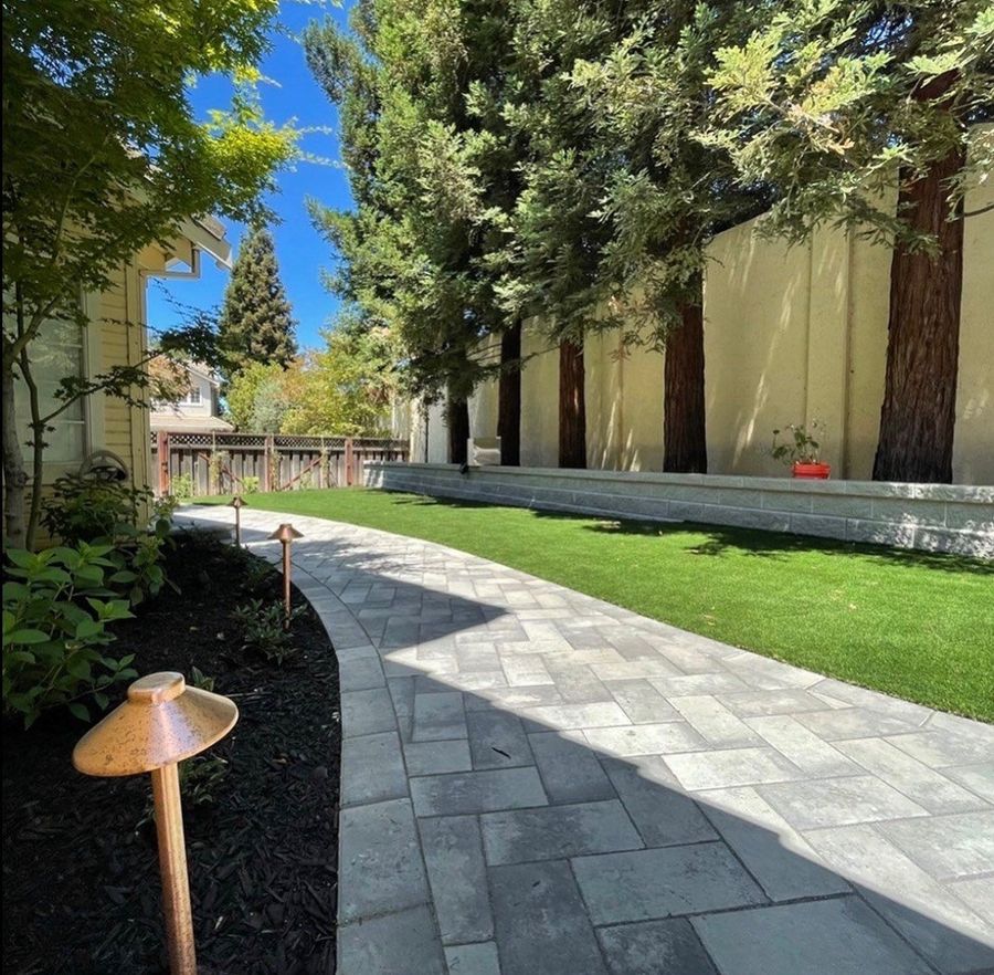 Curved gray paver path with copper path lights beside a dark mulch bed along the house, with a low capped retaining wall on the far side of a green lawn.