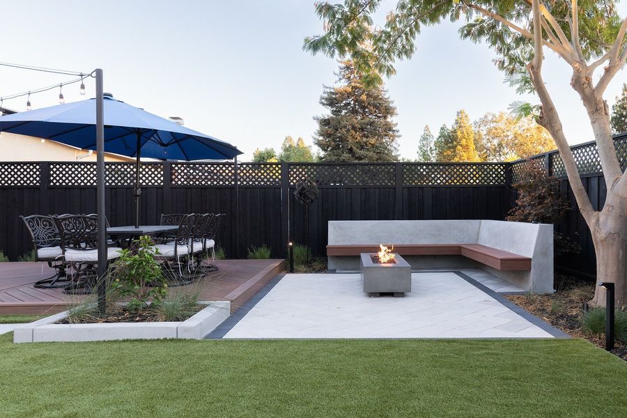 Daytime view across turf to a pale paver court with a fire table, a concrete corner bench and a dining deck under a blue umbrella