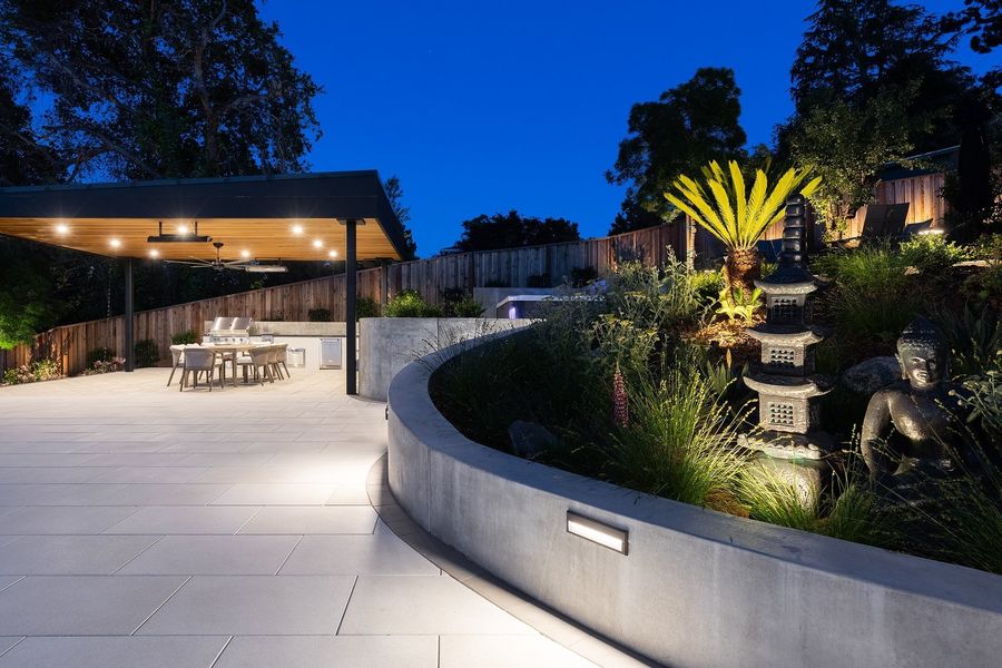 Night view of the curved concrete planter wall with recessed lights, uplit sago palm and stone statuary, and the pavilion beyond