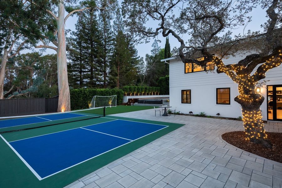 Night view of the white house, an oak tree wrapped in string lights, a blue sports court and grey pavers
