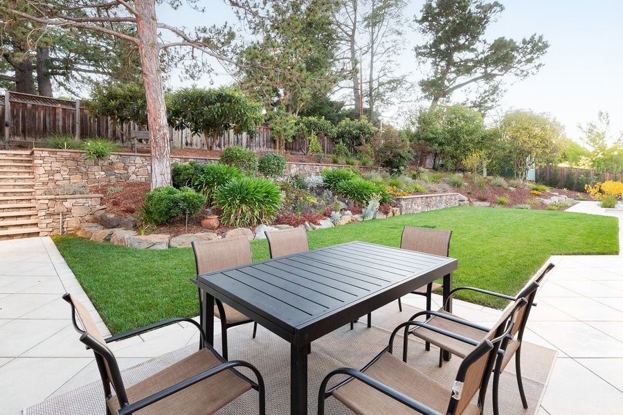 Outdoor dining table at the edge of a lawn on pale paving, with stone terrace walls and layered planting rising behind