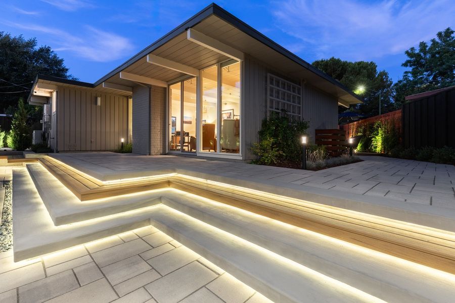 Angled concrete and wood steps outlined in warm strip lighting leading up to the glass walls of the house at dusk