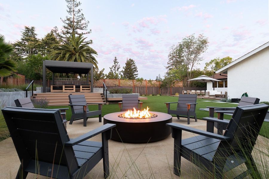 Round concrete patio with a lit gas fire bowl, grey outdoor chairs and the pergola deck in the distance at dusk
