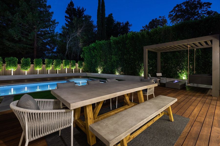 Night view of a concrete dining table and bench on a hardwood deck with an uplit hedge and a pergola swing