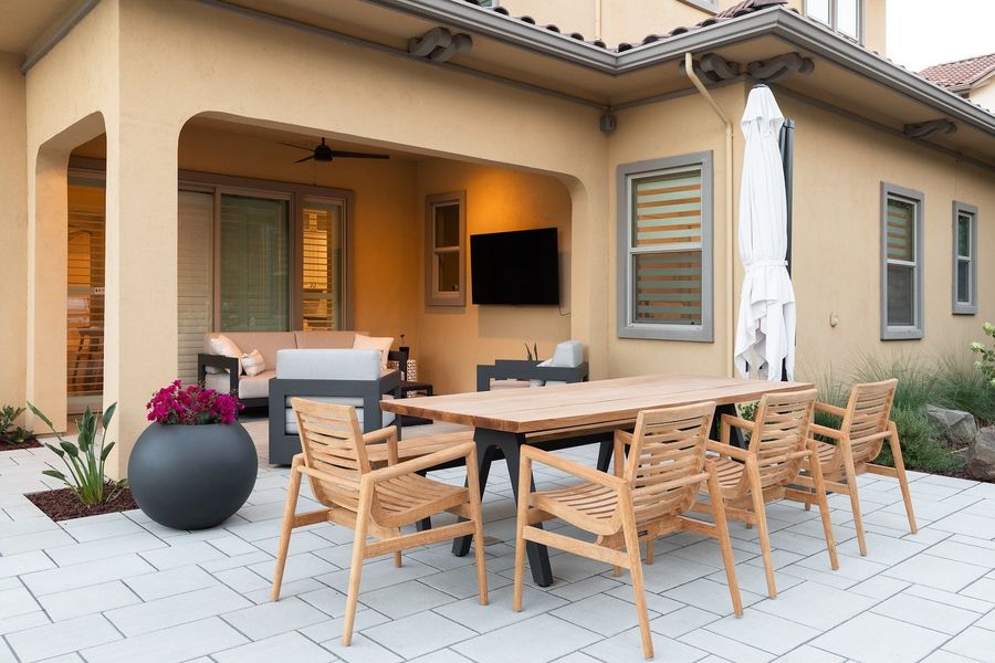Teak dining table and chairs on pavers with a bowl planter of pink flowers in front of the covered patio