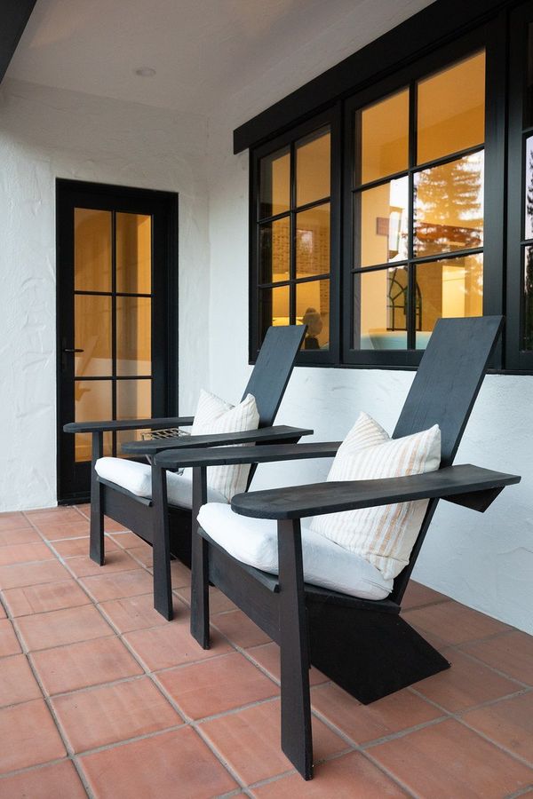 Two dark modern Adirondack chairs with white cushions on a terracotta-tiled porch beside black-framed windows