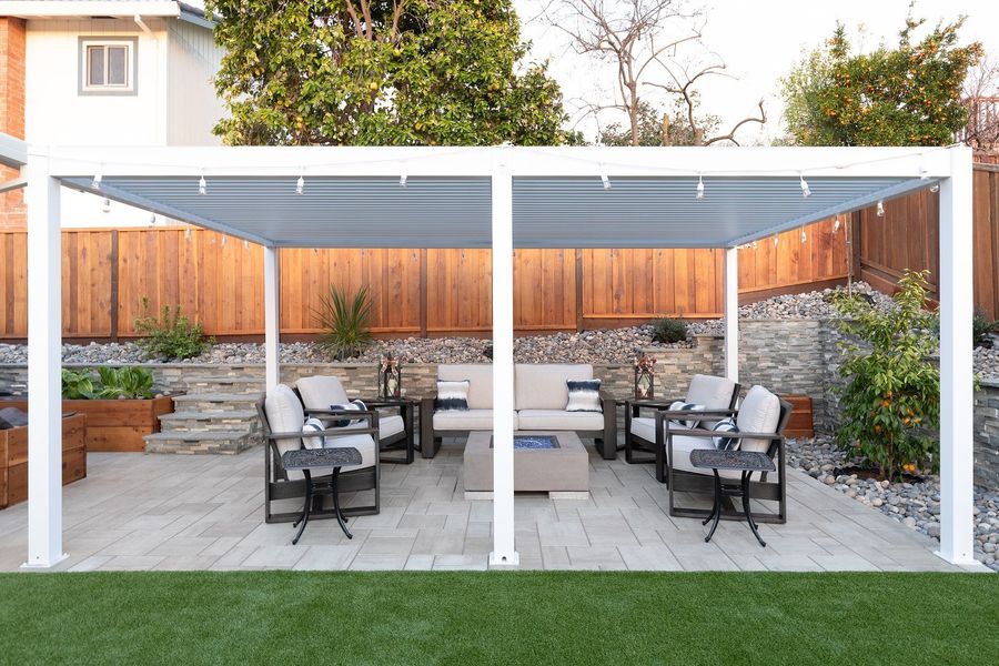 White louvered pergola over a lounge of sofas and a square fire table, with a stacked-stone wall, a wood fence and turf in front