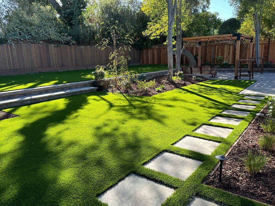 Rectangular concrete stepping pavers crossing a green lawn on a diagonal, with mulch beds, a stepped block wall and a wood pergola in the background.