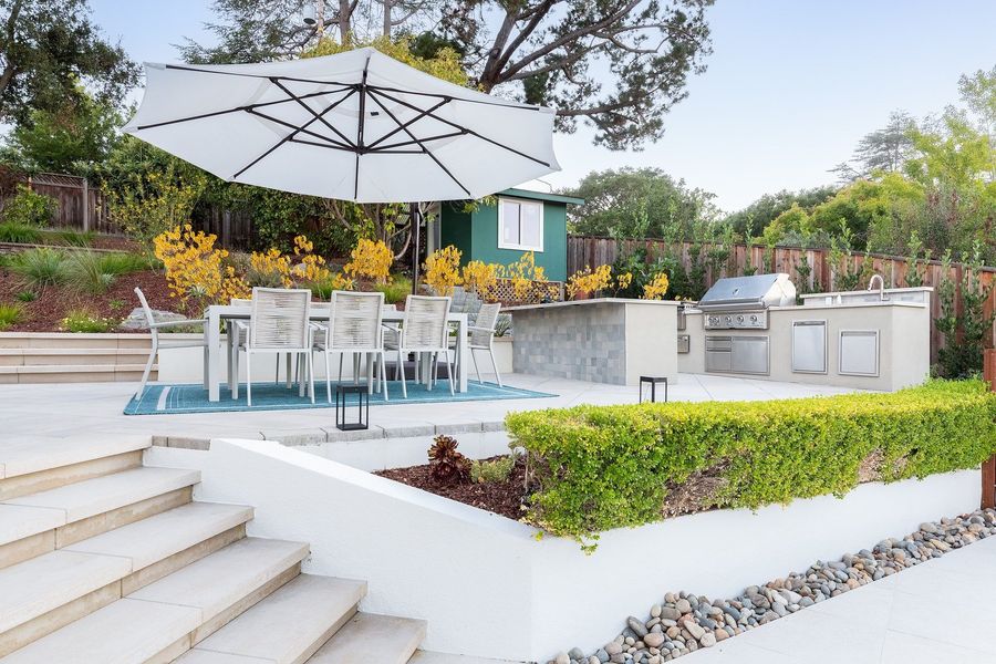Wide view across the terrace to the dining table and kitchen island, with broad steps and a trimmed hedge in front