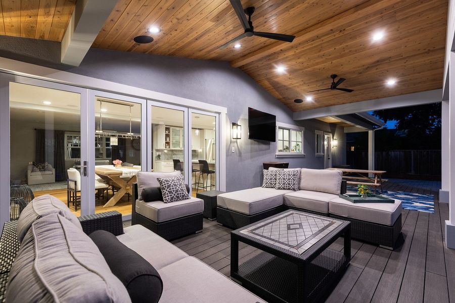 Covered deck with a sectional sofa and lounge chairs under a cedar ceiling with fans and recessed lights, beside folding glass doors and a wall-mounted TV