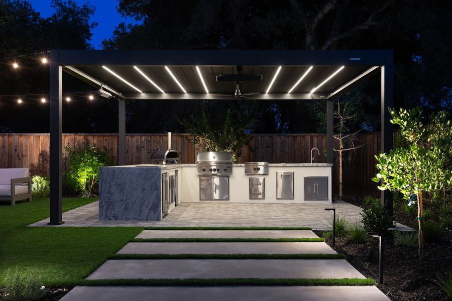 Louvered pergola lit from within over the outdoor kitchen, stone-topped island and built-in grill