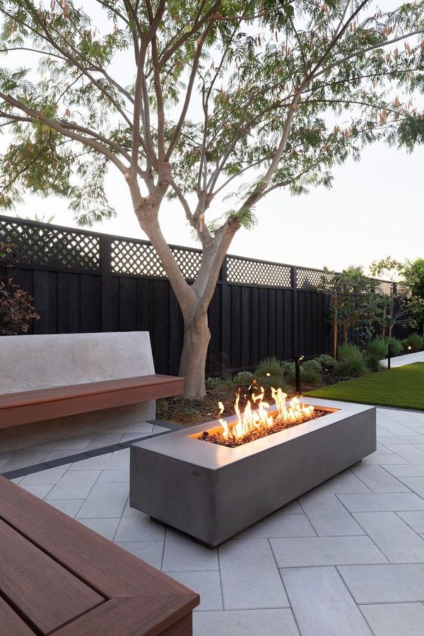 Gas fire table burning on gray pavers beside a wood-seat bench, with a tree and a dark fence behind