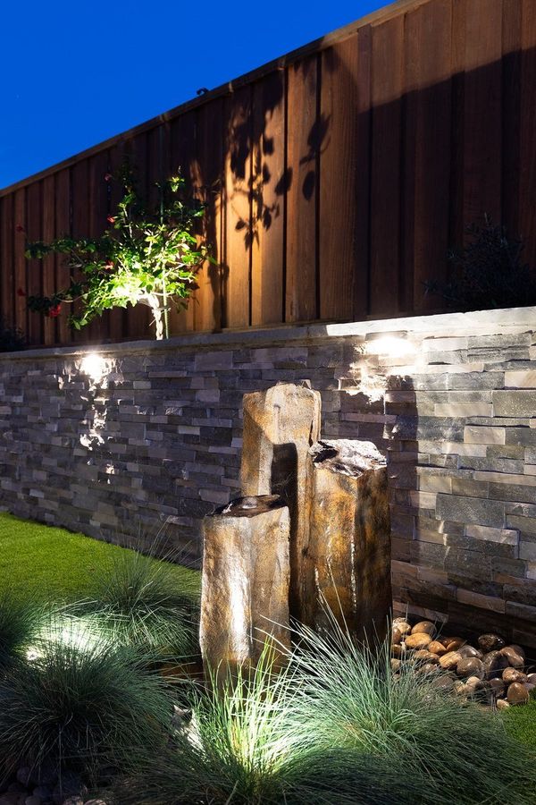 Close night view of three basalt fountain columns against a lit stacked-stone wall with uplit plants and a wood fence above