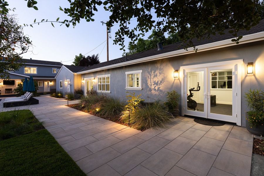 Wide slab paving in front of white French doors, with grasses and shrubs lit by path lights and wall lanterns
