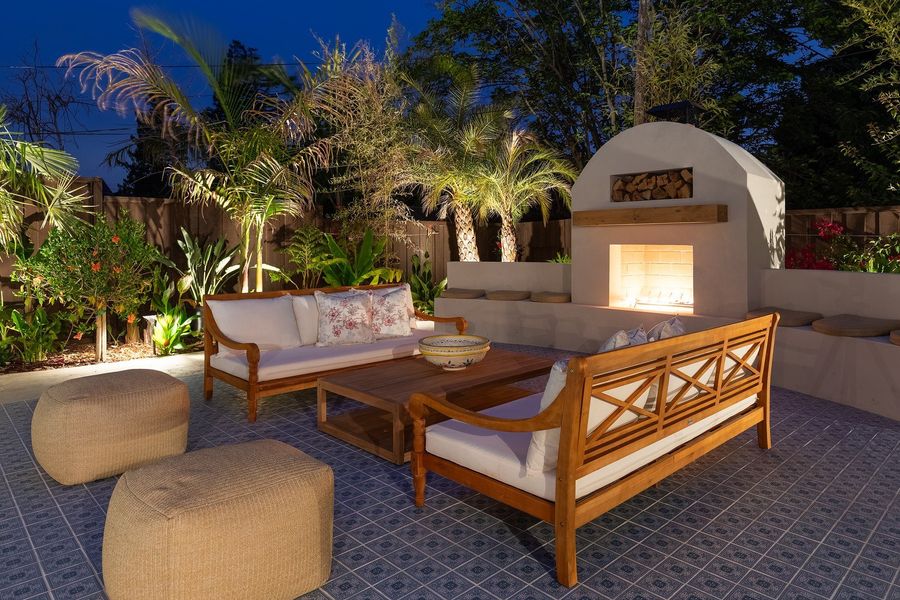 Night view of daybeds, poufs and a glowing stucco fireplace on blue patterned tile surrounded by uplit palms