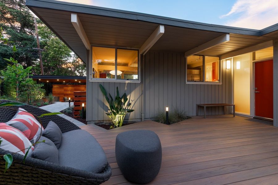 Wood-look deck in front of a mid-century home with a red front door, a lounge chair and uplit leafy plants